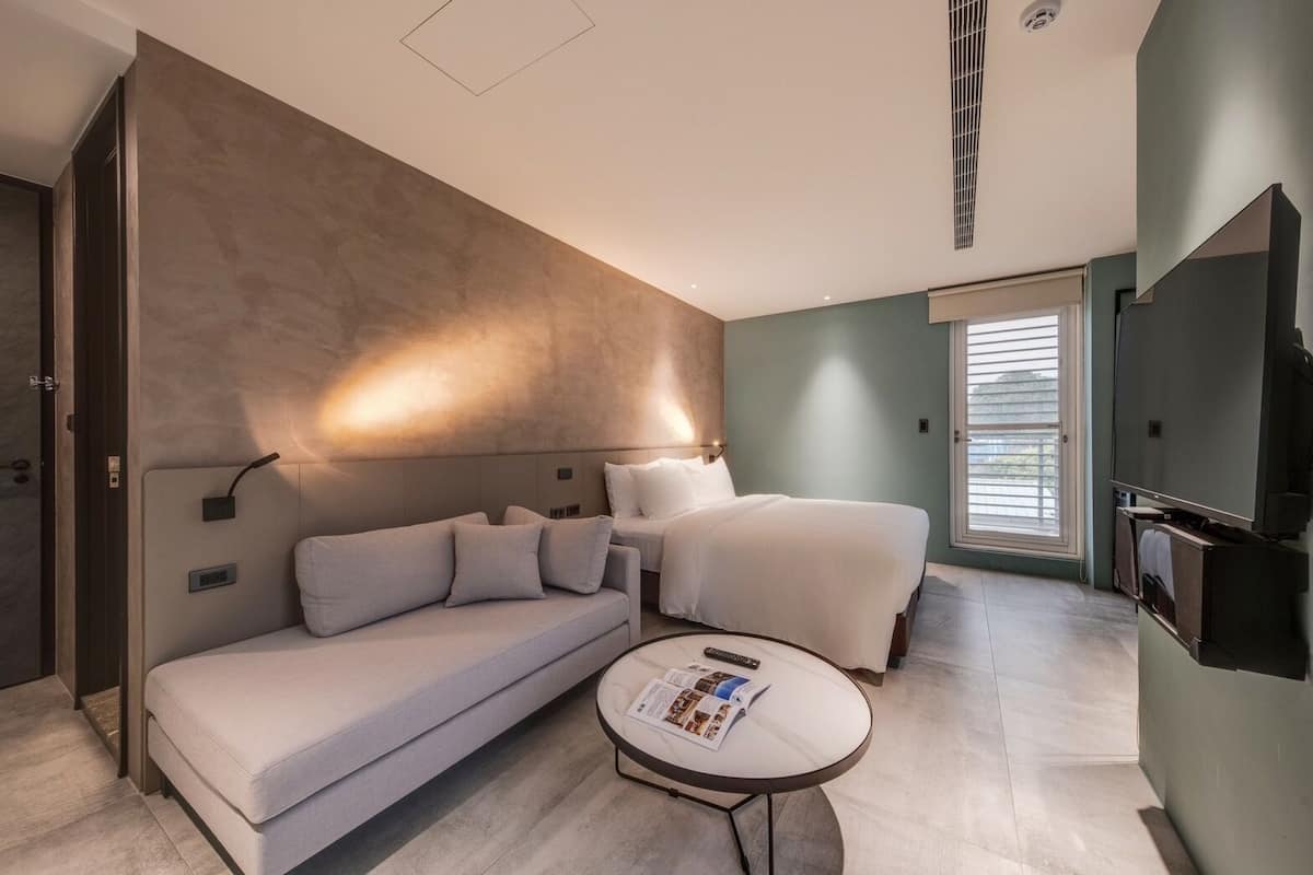 Ein modernes Hotelzimmer mit einer Couch, einem Bett, einem kleinen runden Tisch mit einer Zeitschrift und einem Fernseher.