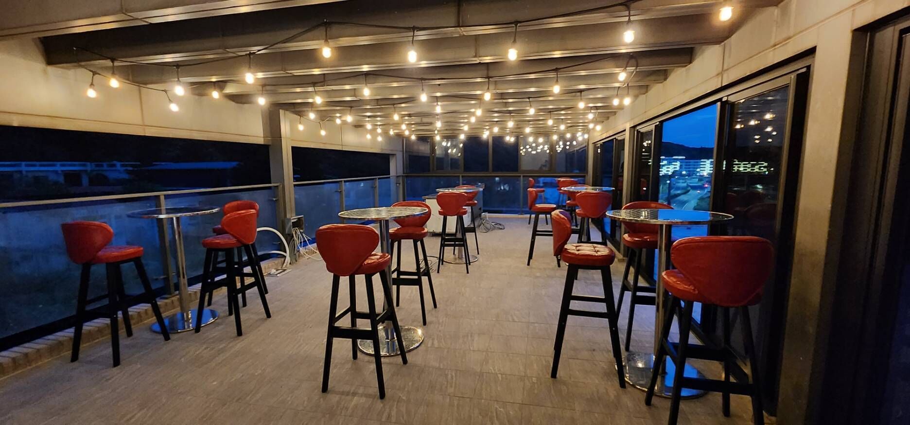 A covered outdoor seating area with red chairs and tables, large windows offering a view of the cityscape.