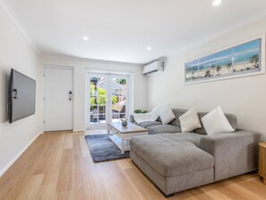 A modern living room with a grey sectional sofa, a flat-screen TV mounted on the wall, and a large window with a view of the outdoors.