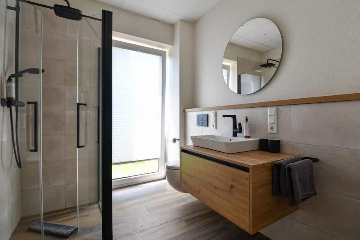 A modern bathroom with a glass shower enclosure, a wooden vanity, a round mirror, and a view of the outdoors.