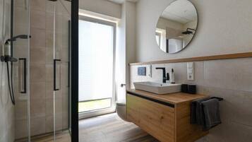 Een moderne badkamer met een glazen douchecabine, een houten wastafelmeubel, een ronde spiegel en uitzicht op de natuur.