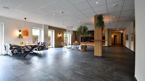 Un hall d’hôtel spacieux et moderne, doté d’un plafond haut, d’un sol carrelé foncé et de différents espaces de détente, notamment une table à manger avec des chaises et un coin salon avec des canapés.