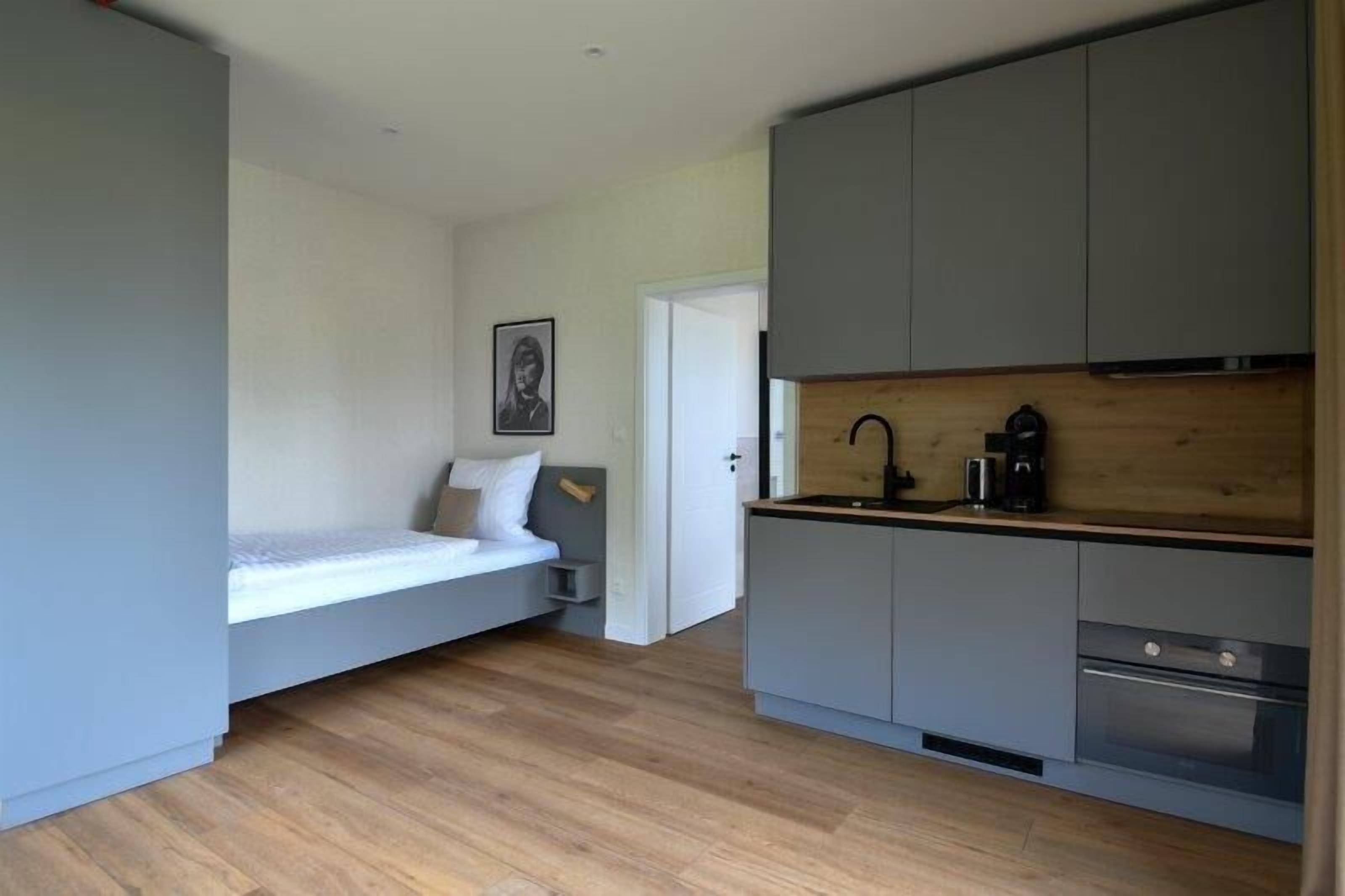 A compact room with a bed, a kitchenette, and a door leading to another room.