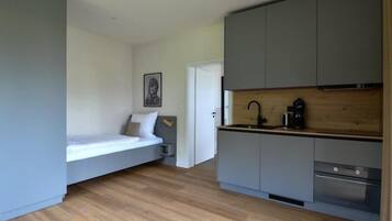 A compact room with a bed, a kitchenette, and a door leading to another room.