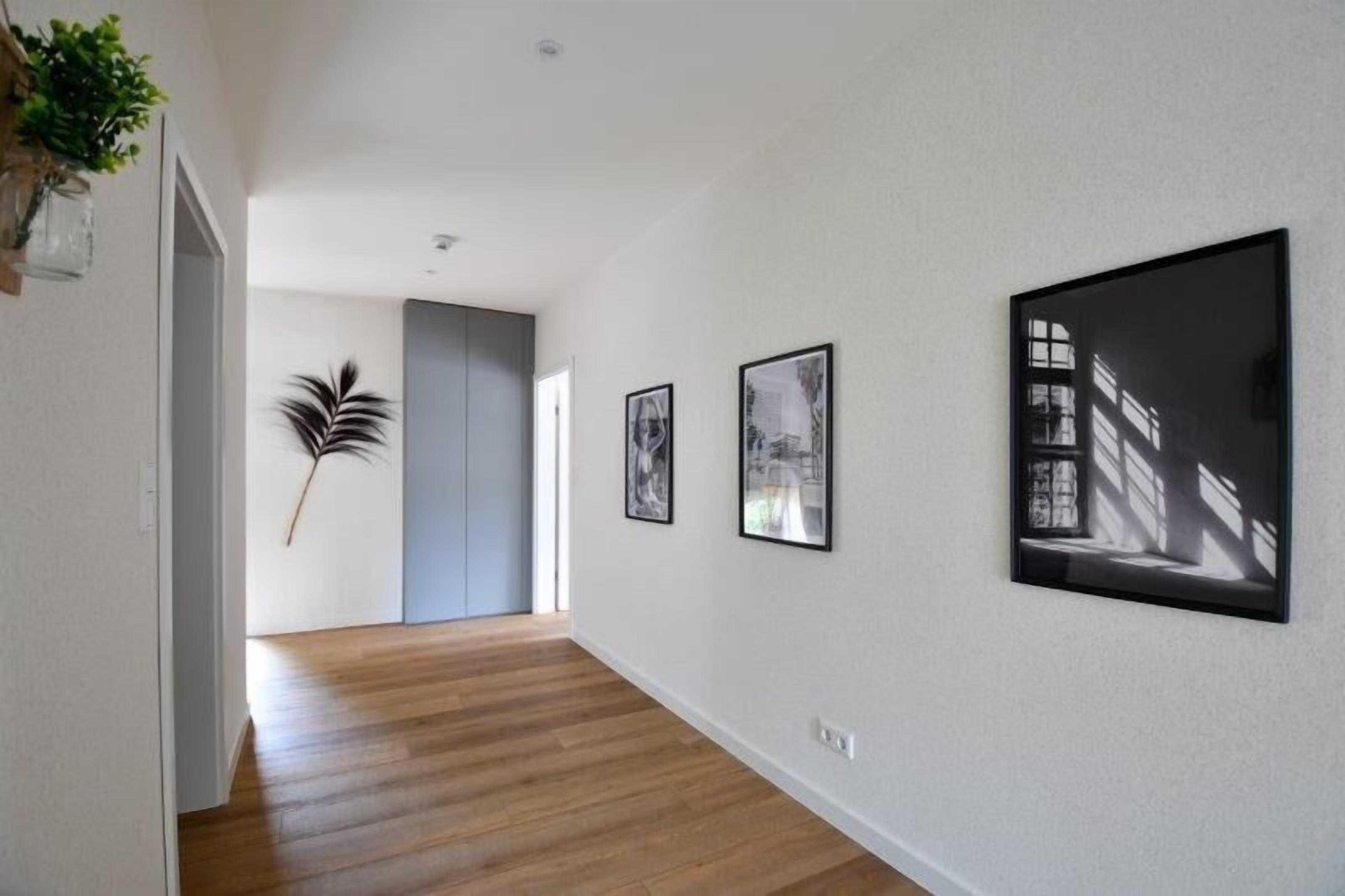 A hallway with wooden flooring, a plant on the left, and three framed pictures on the right wall.