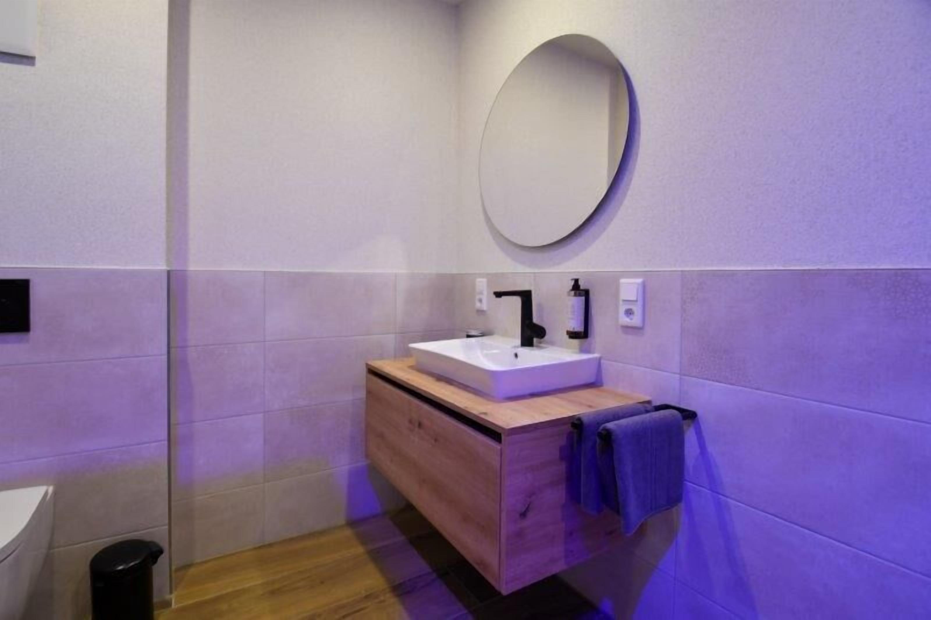 Een moderne badkamer met een witte wasbak, een houten opbergmeubel en een ronde spiegel.