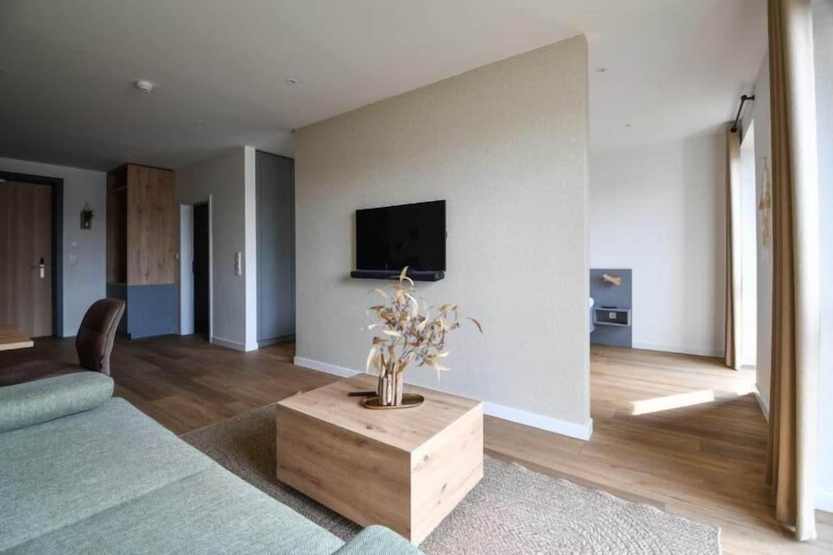 A modern living room with a wooden coffee table, a flat-screen TV mounted on the wall, and a large window providing natural light.