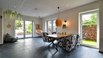 A modern dining area with a wooden table and floral chairs, large windows, and hanging plants.