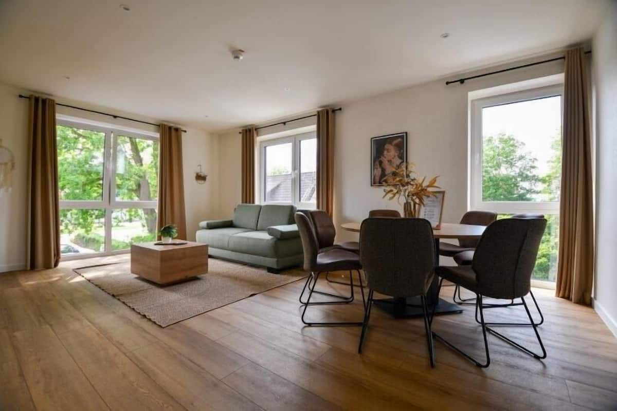 A spacious room with a wooden floor, a green sofa, a dining table with chairs, and large windows with curtains.