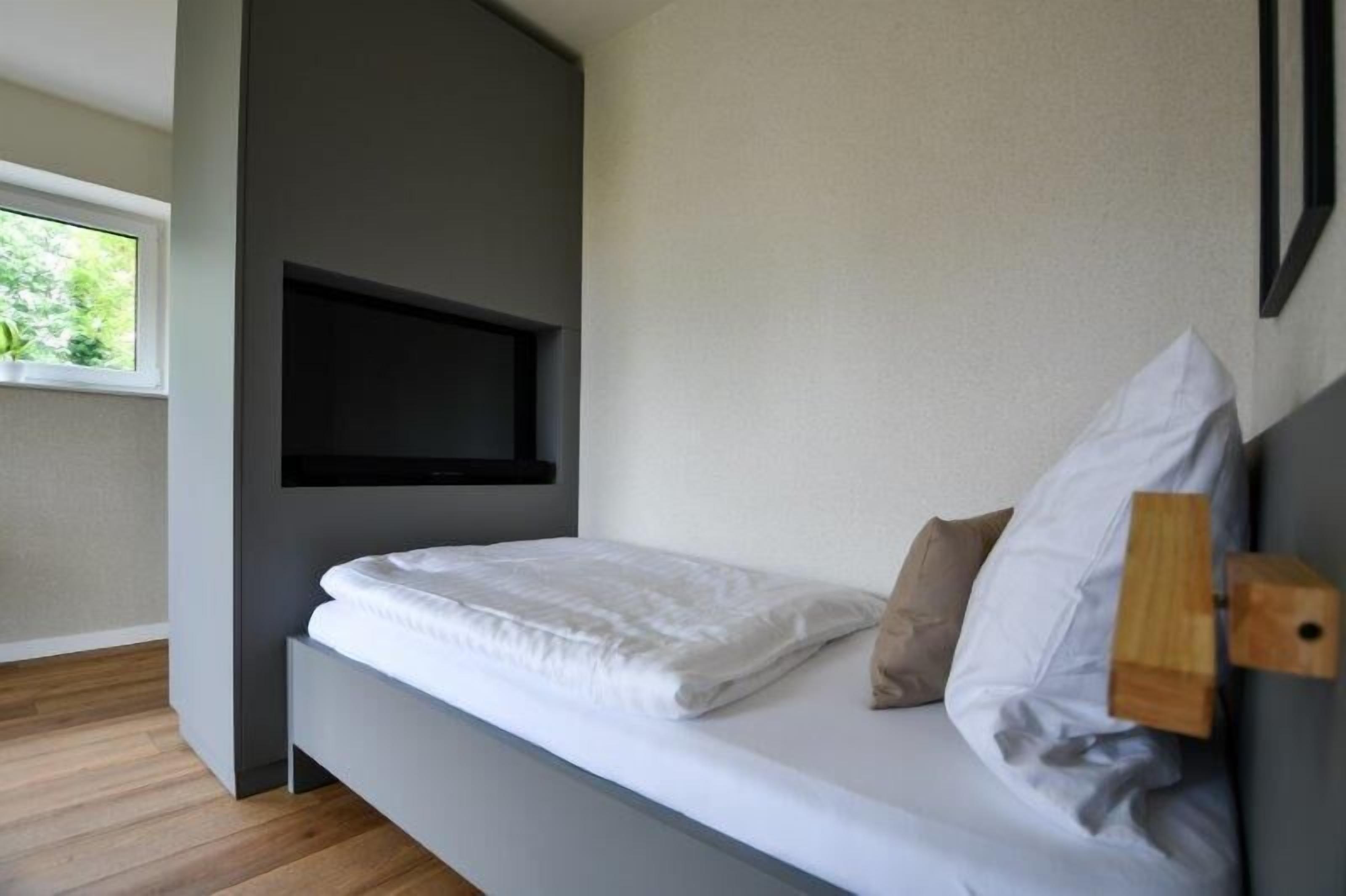 A modern bedroom with a bed, a mounted TV, and a window with a view of greenery.