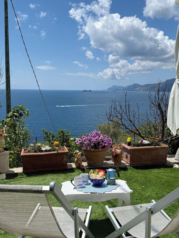Une terrasse avec une table dressée pour deux personnes, entourée de plantes en pot et offrant une vue panoramique sur la mer et les îles lointaines.
