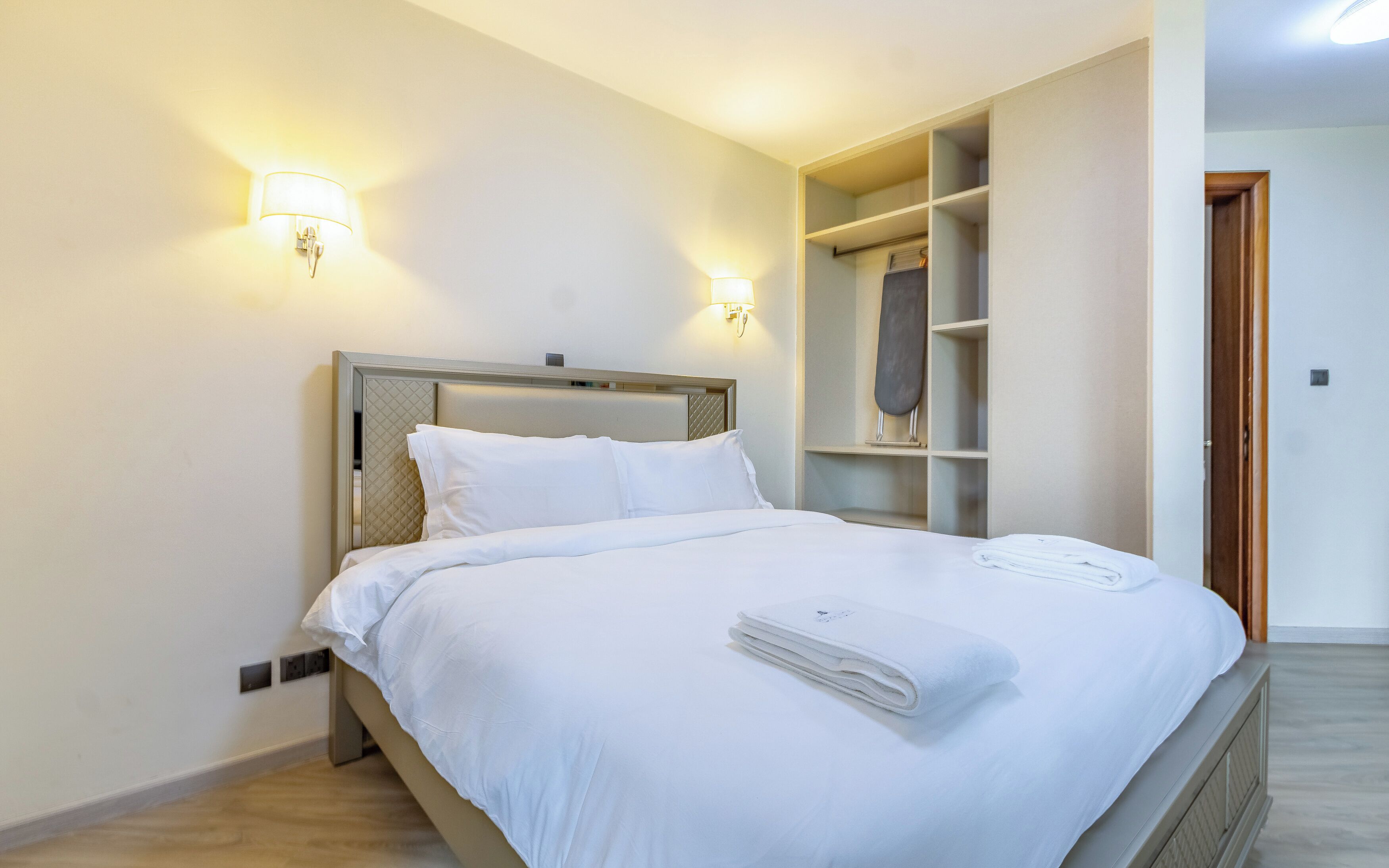 Een net opgemaakt bed met witte lakens en een gevouwen handdoek, een hoofdbord met een structuur en twee wandlampen.