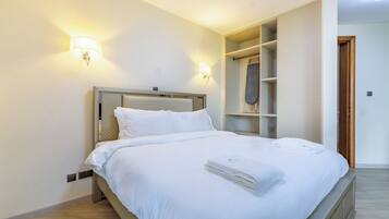 Een net opgemaakt bed met witte lakens en een gevouwen handdoek, een hoofdbord met een structuur en twee wandlampen.