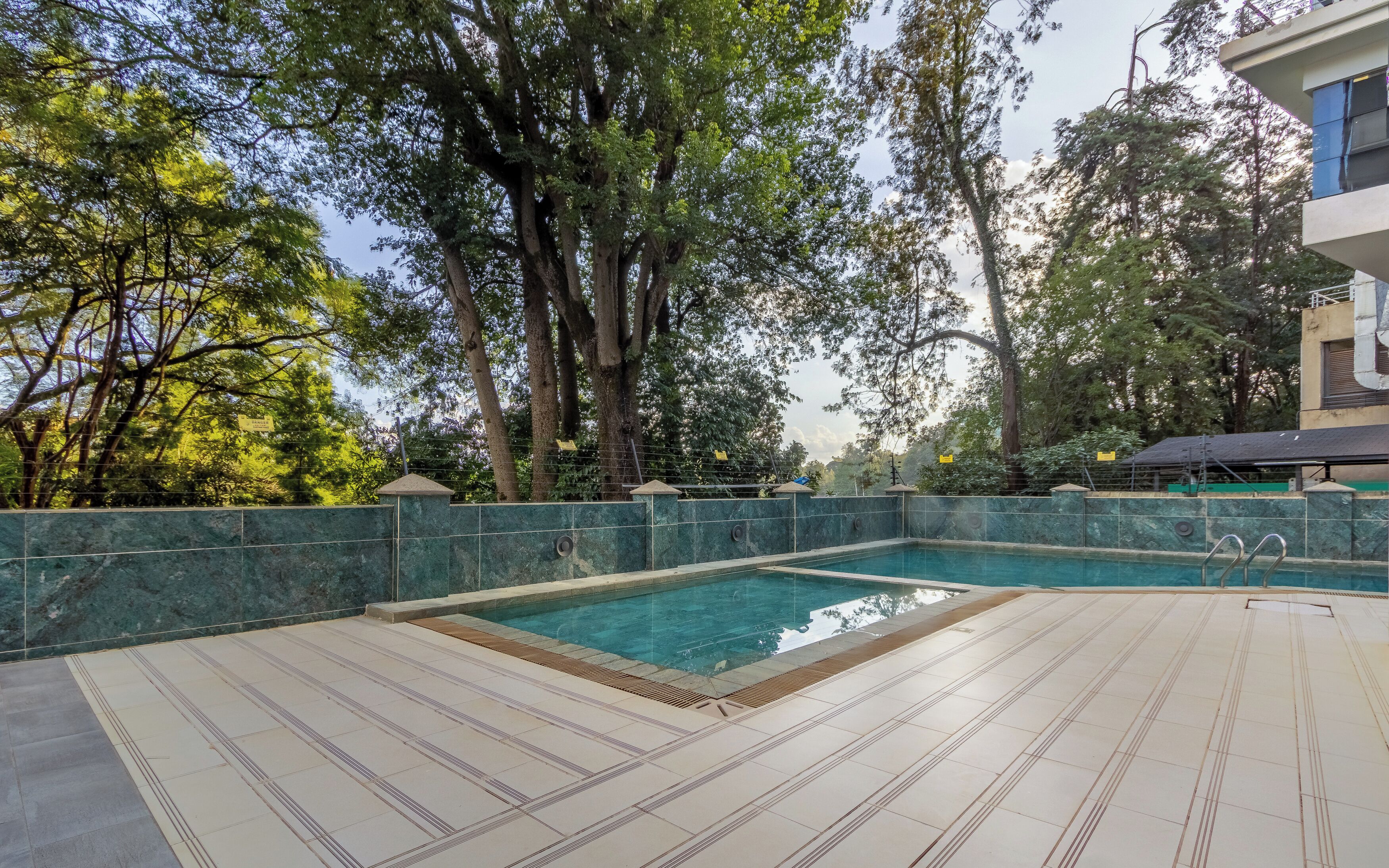 A swimming pool with a tiled deck, surrounded by trees and a building.
