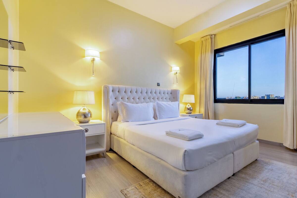 A hotel room with a large bed, two bedside lamps, a nightstand, a window with curtains, and a view of the cityscape.