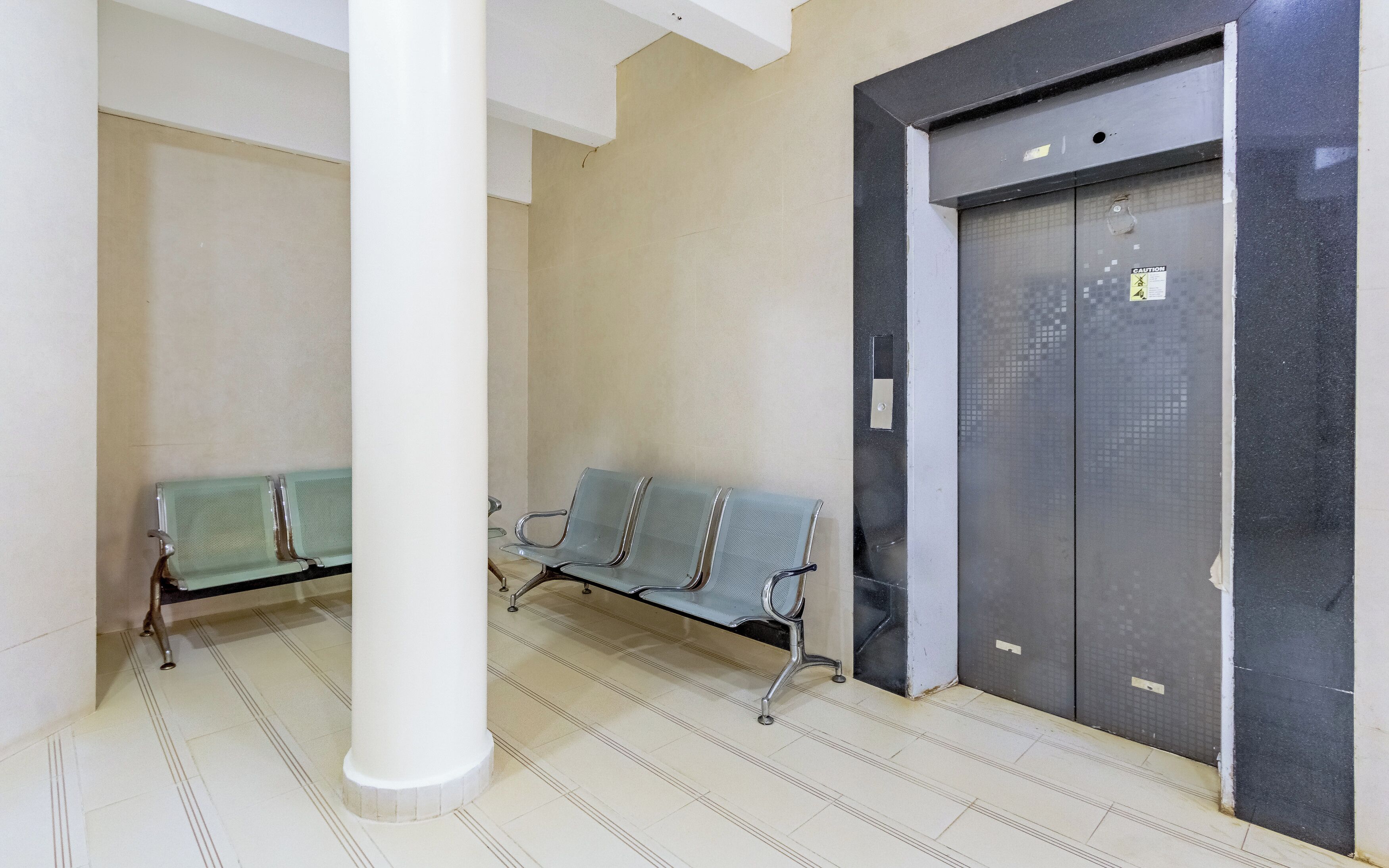 A waiting area with two metal benches and a pillar, an elevator with caution tape, and tiled flooring.
