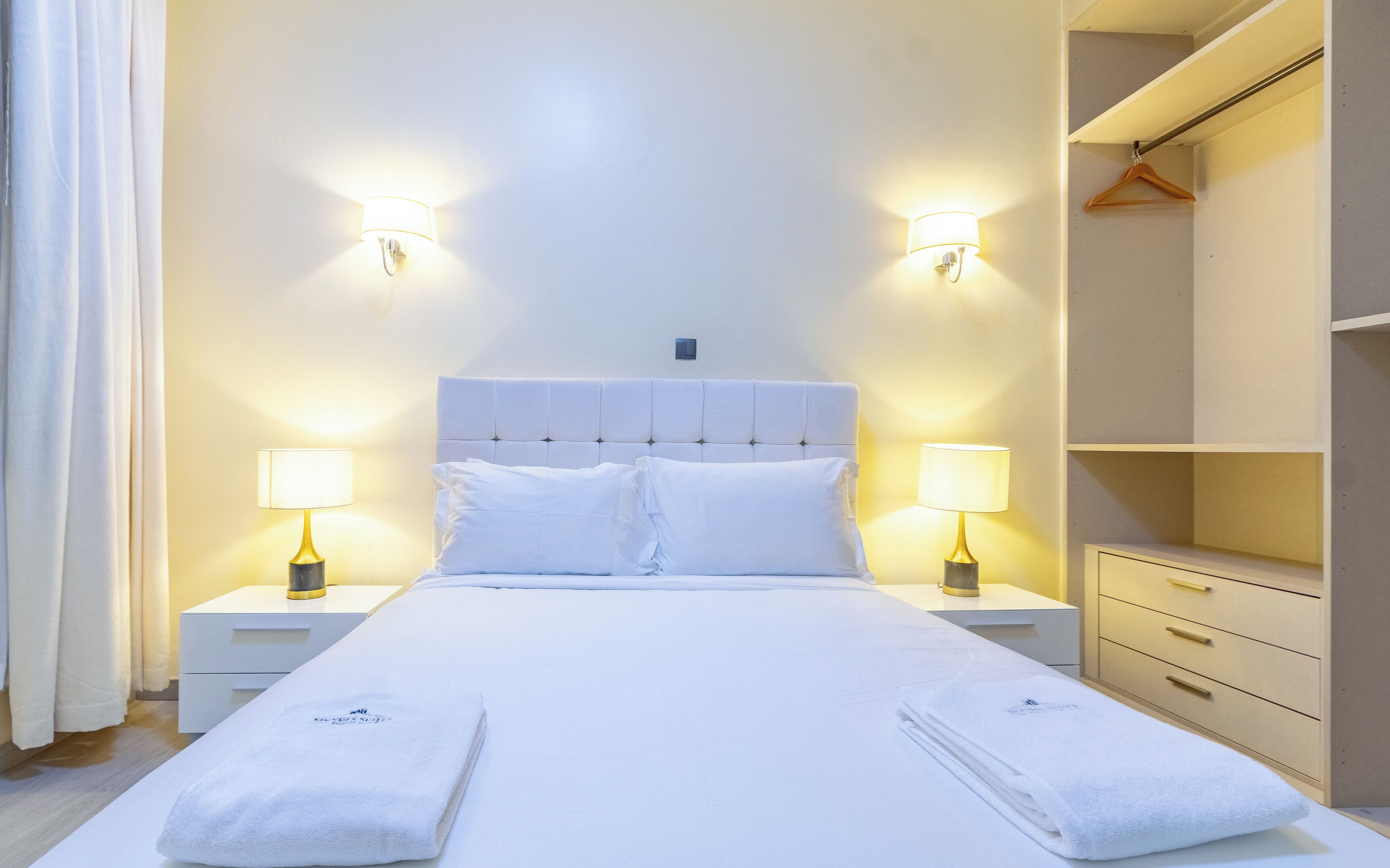 A neatly made bed with two pillows, two bedside lamps, a bedside table, and a wardrobe with a hanging rack.