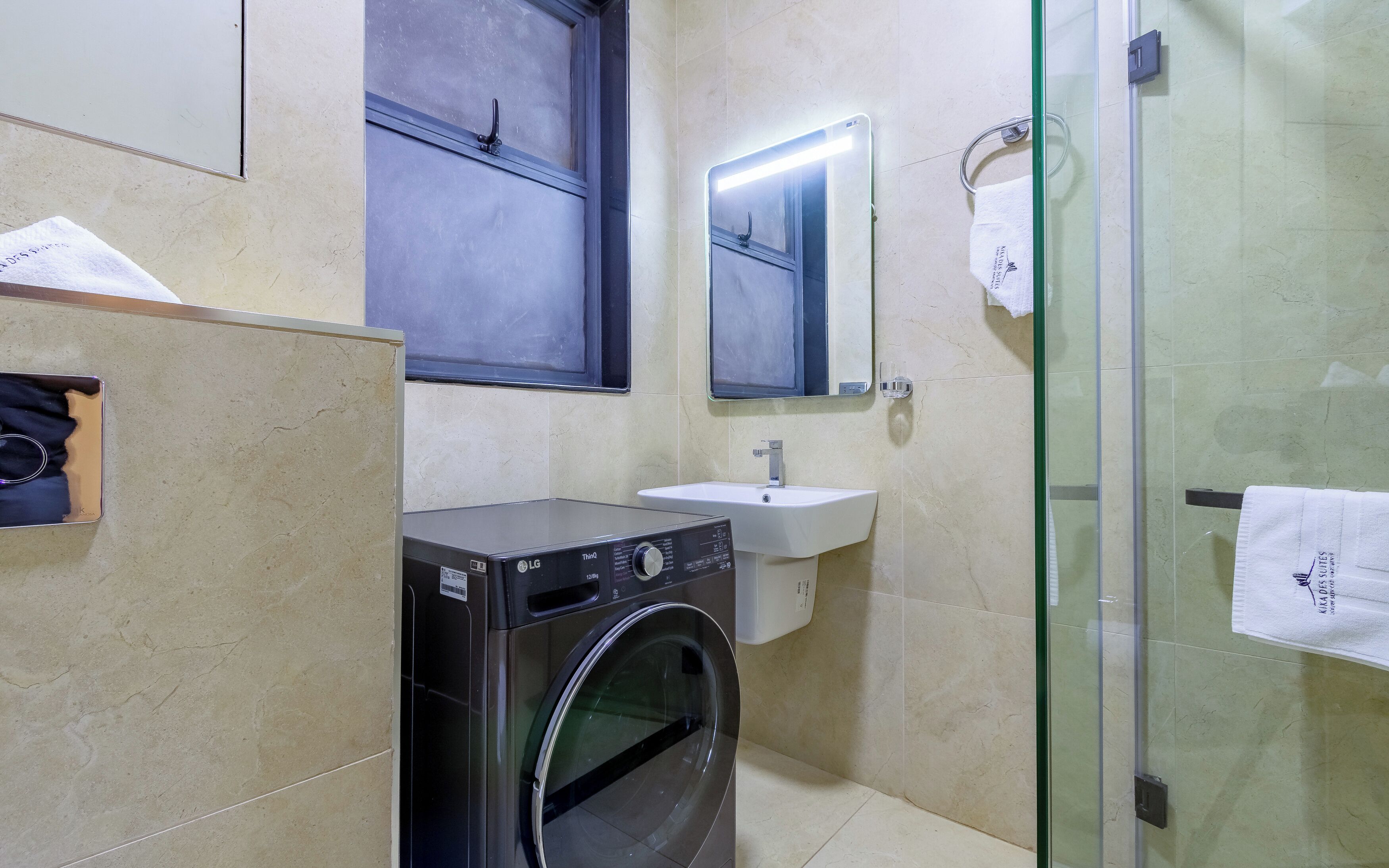 A compact hotel bathroom with a washing machine, a sink, a glass shower enclosure, and a mirror.
