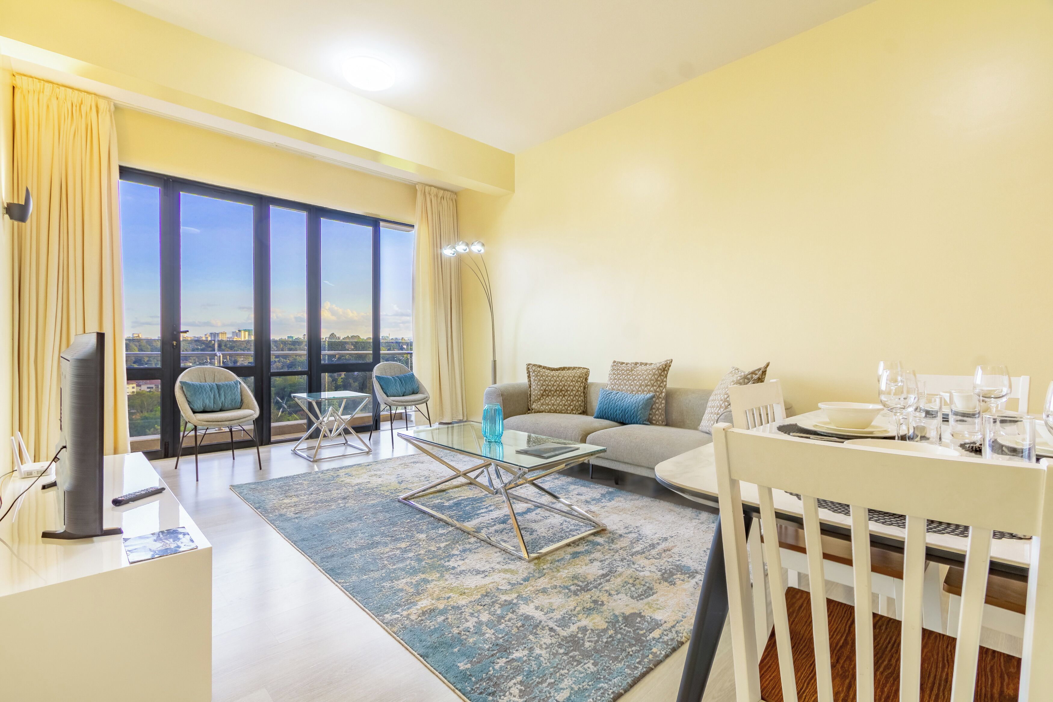 Ein modernes Wohnzimmer mit Sofa, Glastisch, Esstisch und großem Fenster mit Blick auf die Stadt.