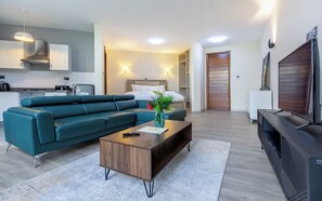 A modern hotel room with a teal sofa, a wooden coffee table, a TV, a kitchen area, and a bed with white linens. - Kika Des Suites - Westland, Nairobi (Nairobi)