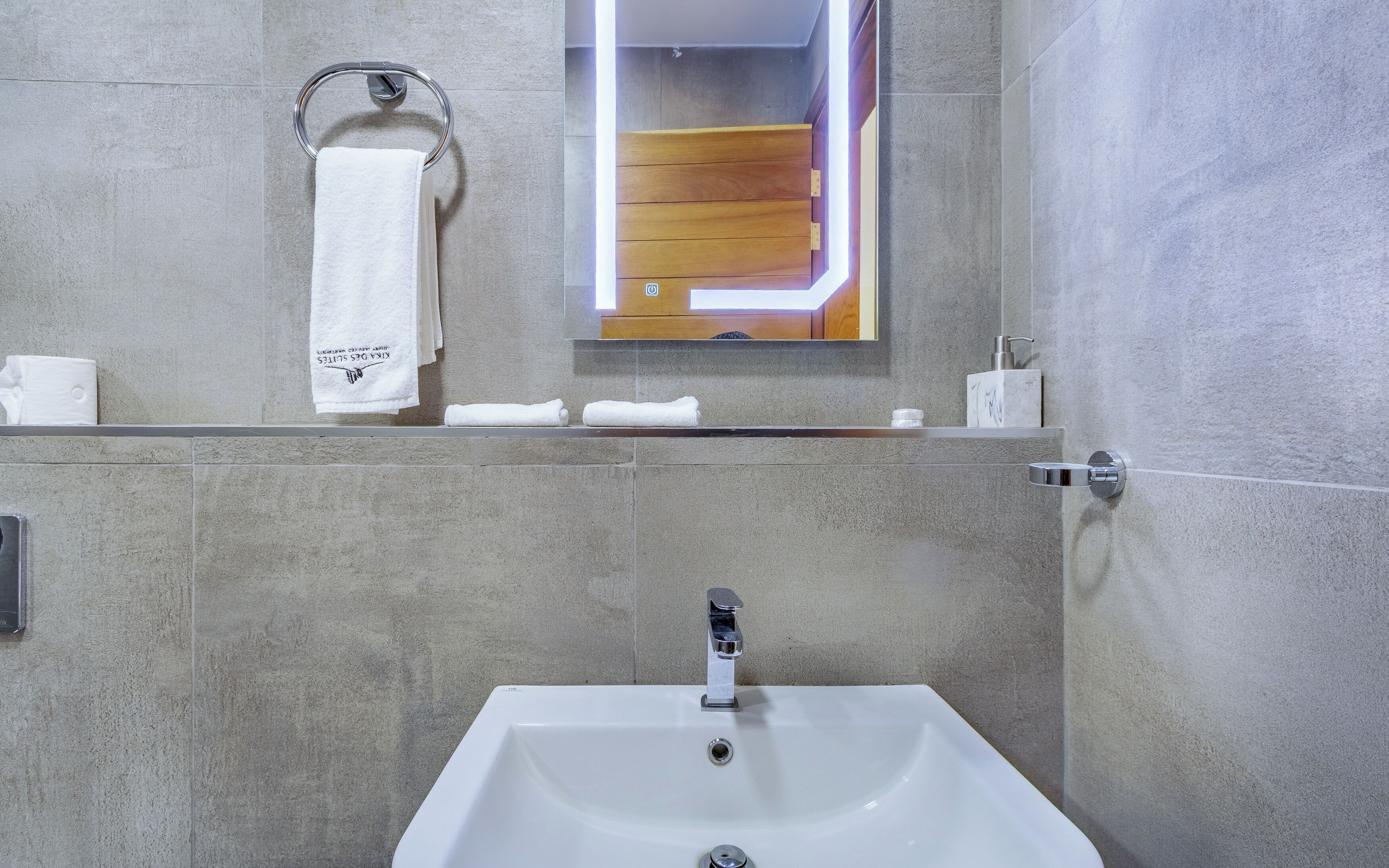 A bathroom with a white sink, a towel rack with a white towel, a mirror, and a wooden panel above the mirror.