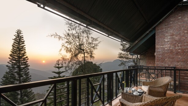 Un balcón con mobiliario de mimbre y vistas a la puesta de sol sobre un paisaje montañoso.