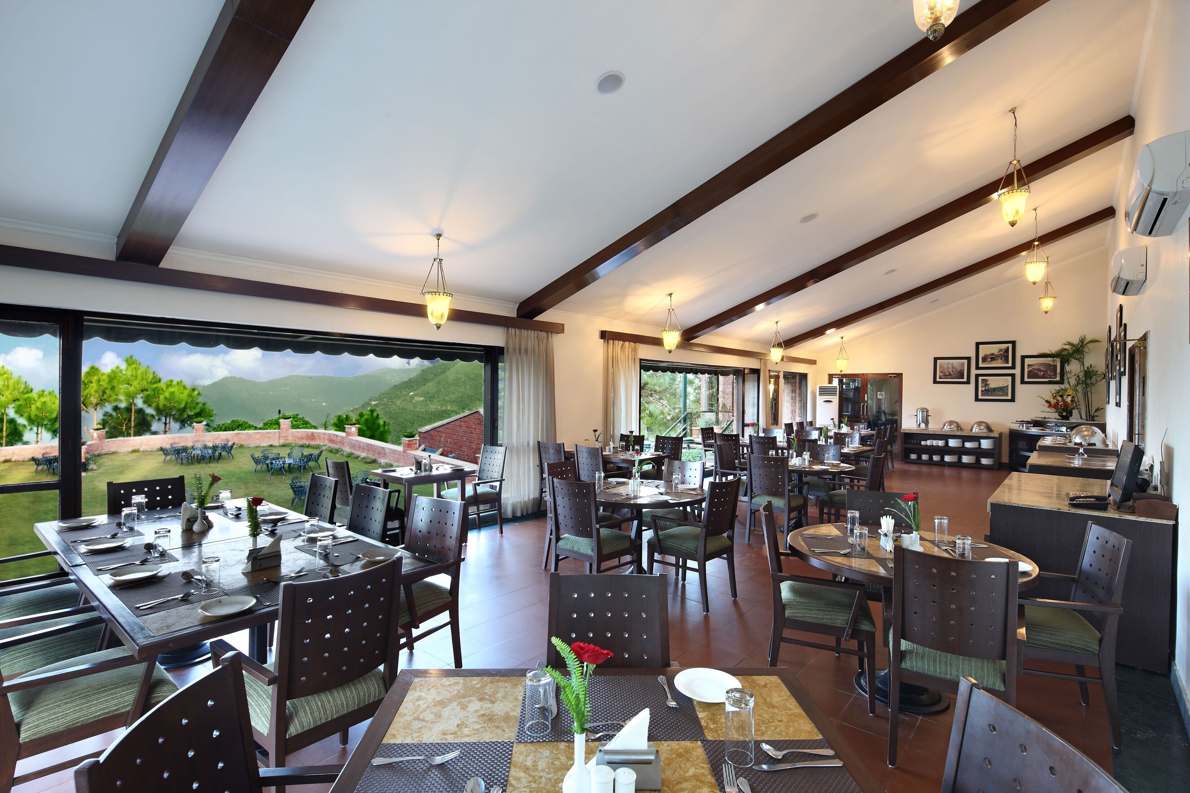 A spacious dining area with multiple tables and chairs, large windows offering a view of the outdoors, and a modern ceiling design.