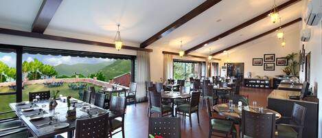 A spacious dining area with multiple tables and chairs, large windows offering a view of the outdoors, and a modern ceiling design.
