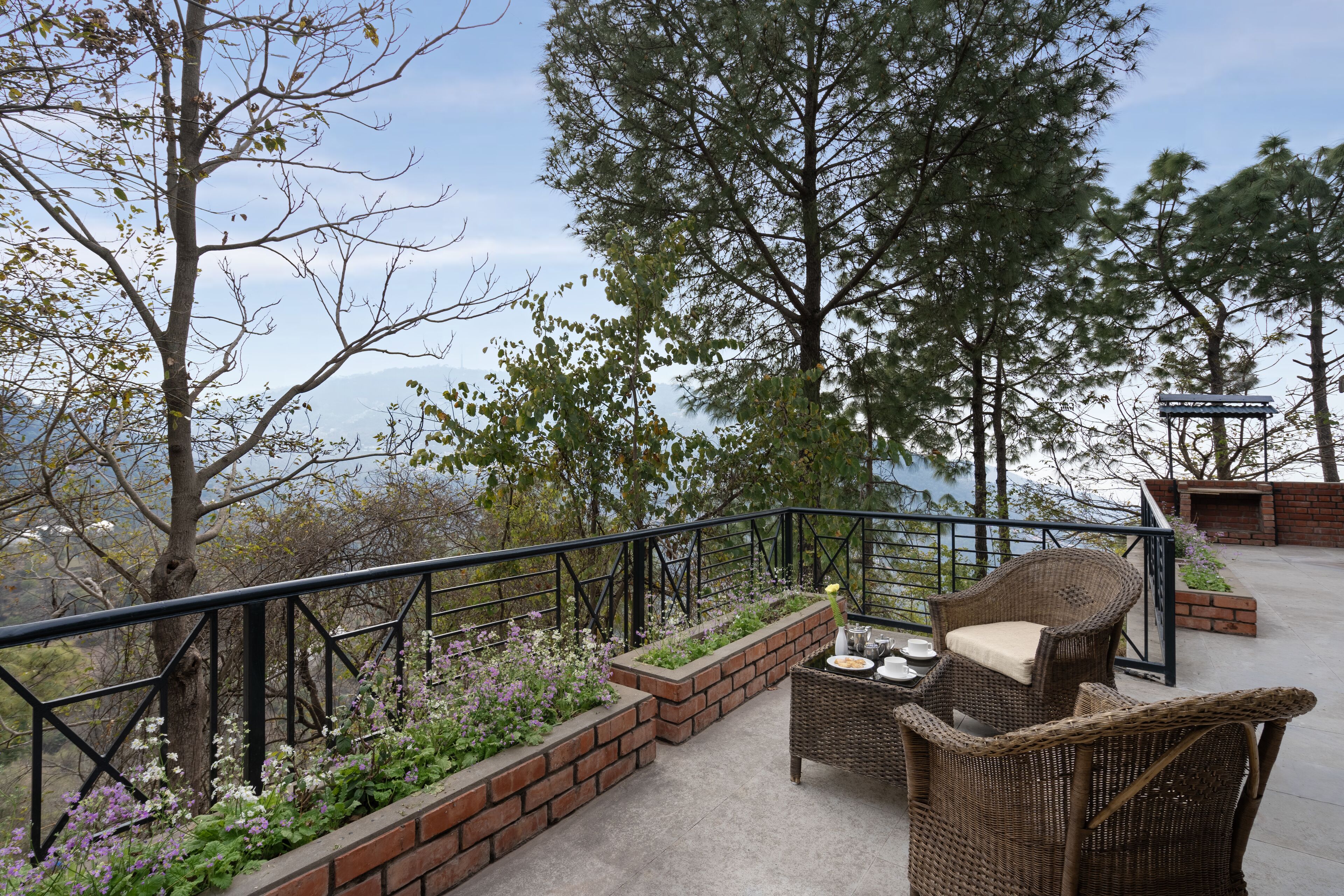 A balcony with wicker furniture, brick planters with flowers, and a view of trees and a distant landscape.