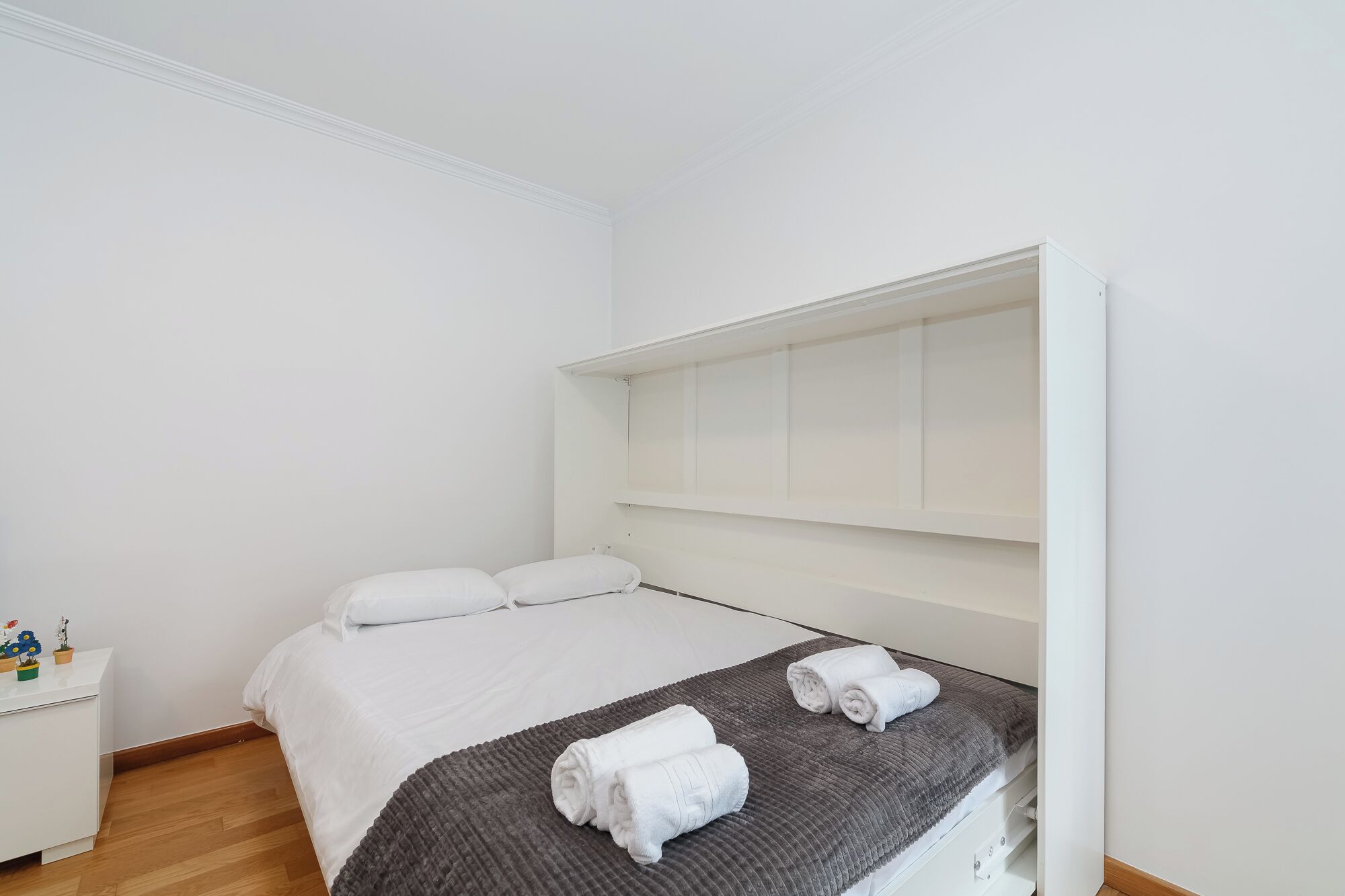 Een minimalistische slaapkamer met een enkel bed, wit beddengoed, een grijze deken en twee opgerolde handdoeken.