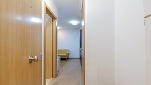 Ein schmaler Flur mit Holztüren links, einem beigen Sofa im Hintergrund und einer weißen Wand.