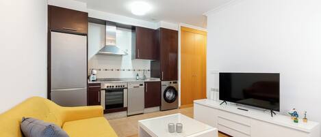 Un espacio habitable compacto con sofá, una mesa de centro, televisión, lavadora y zona de cocina.