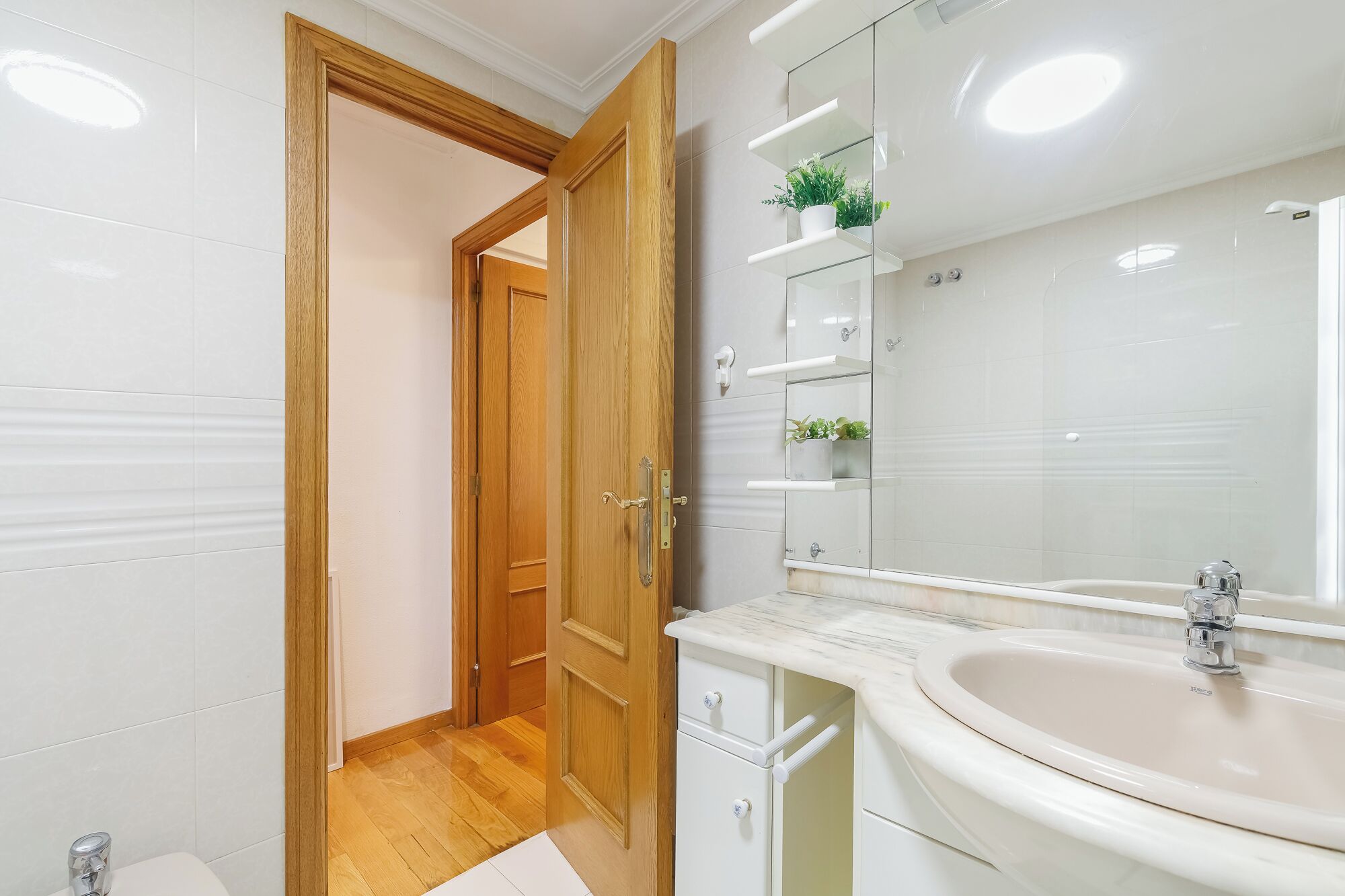 Une salle de bain avec une porte en bois, des carreaux blancs, un lavabo en marbre et une étagère en verre ornée de plantes.