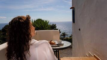 Una persona sentada a una mesa con un croissant y una taza, con vista a un paisaje marino y vegetación.