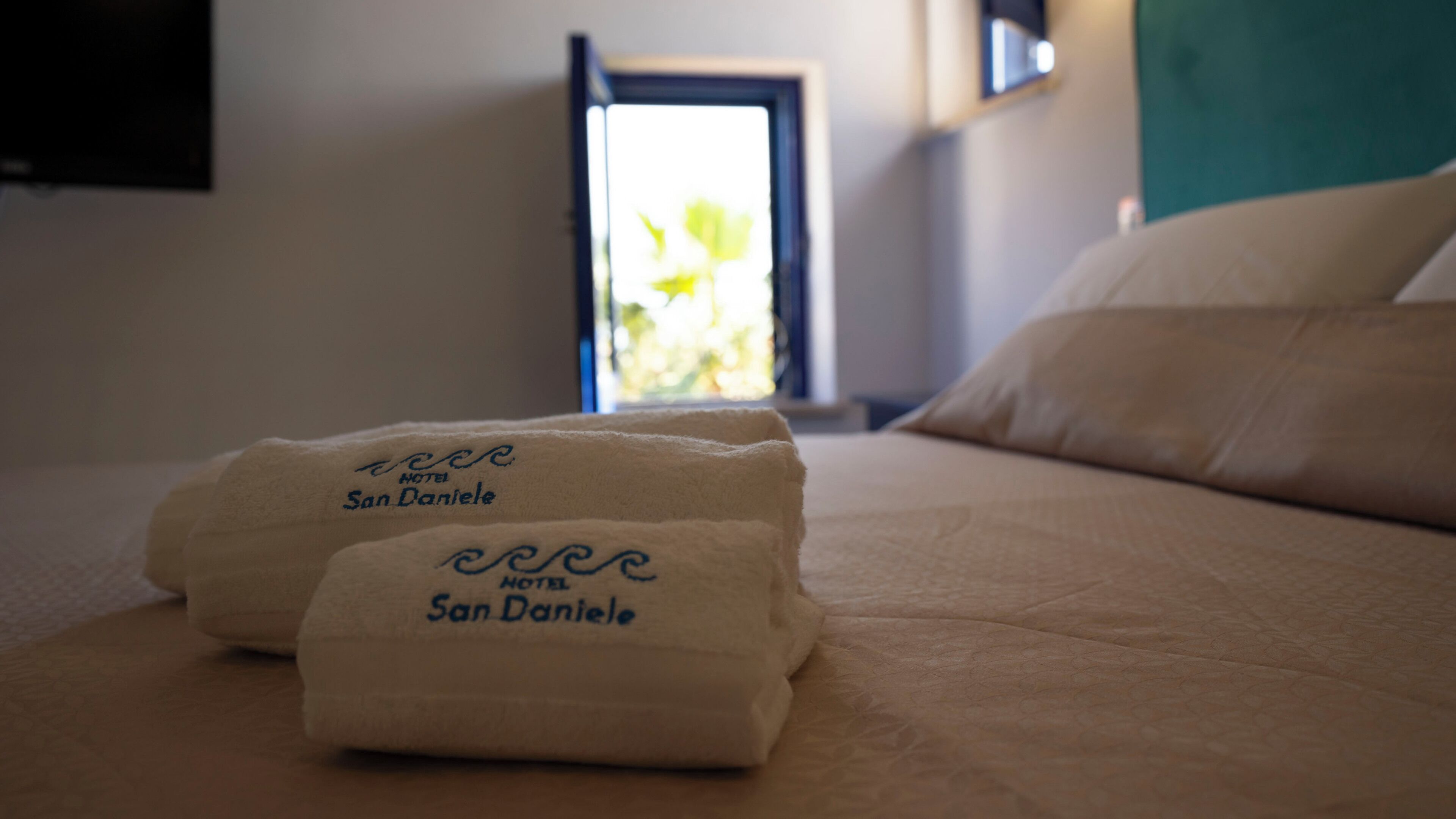 A bed with two towels labeled "Hotel San Daniele" and a window with a view of greenery.