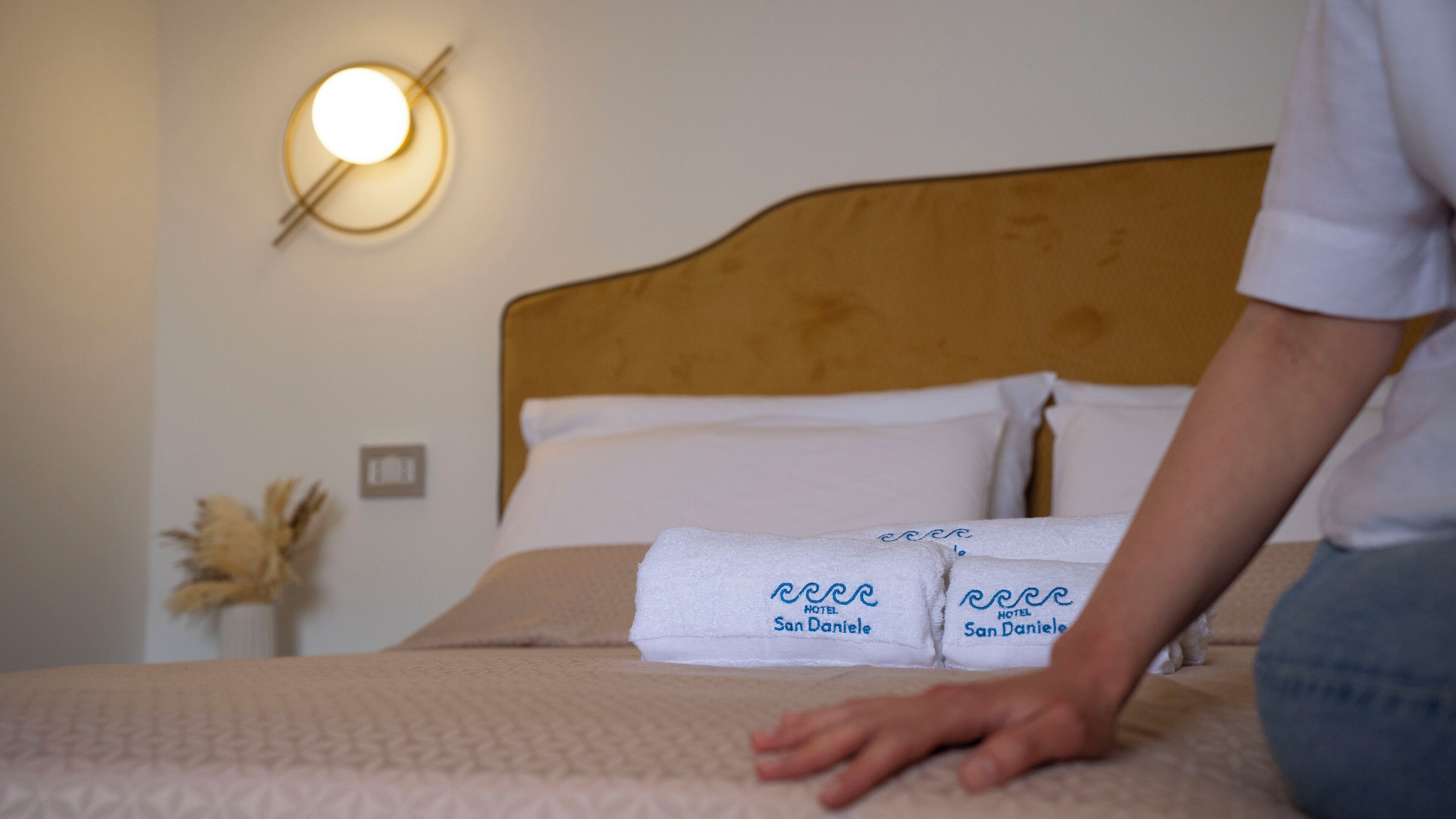 Cama bem arrumada com duas toalhas com a etiqueta "HOTEL San Daniele" e um aplique de luz na parede.