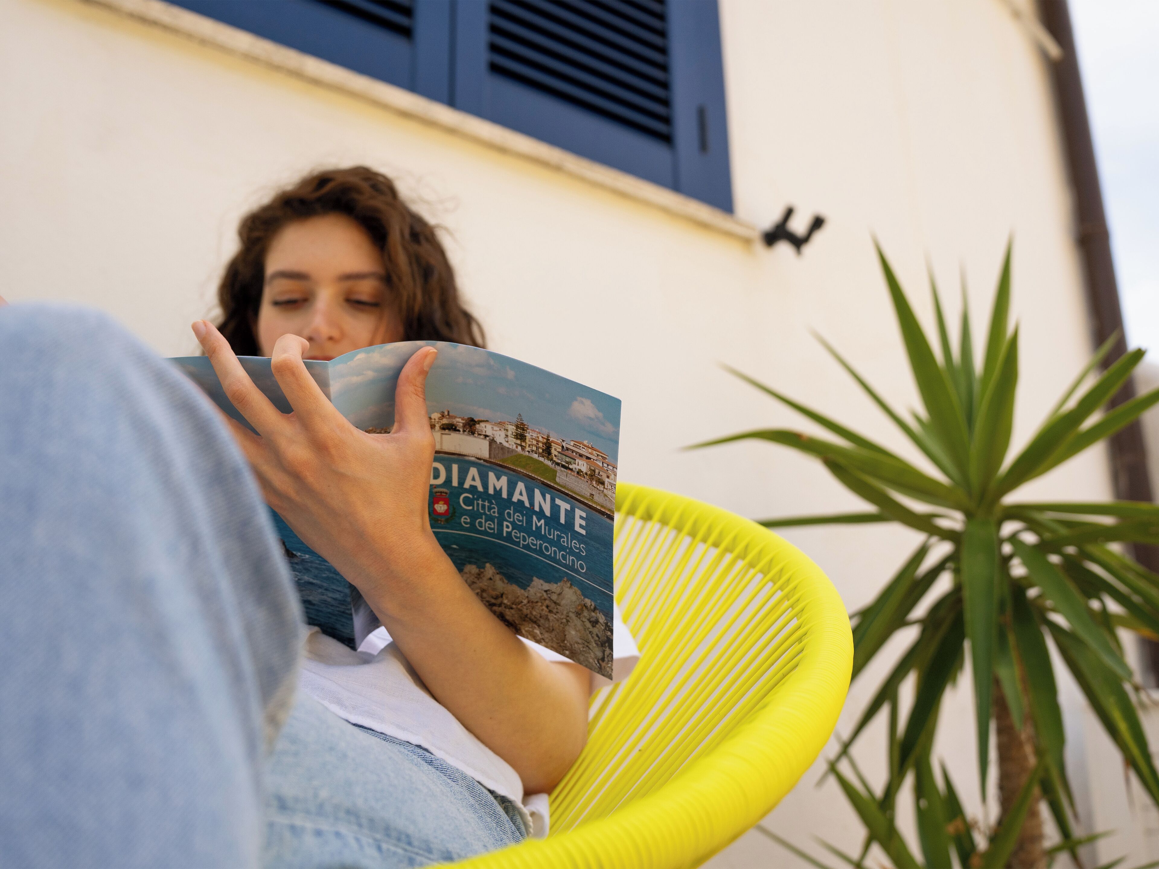 A person reading a book titled "DIAMANTE" in a yellow chair with a plant beside them.