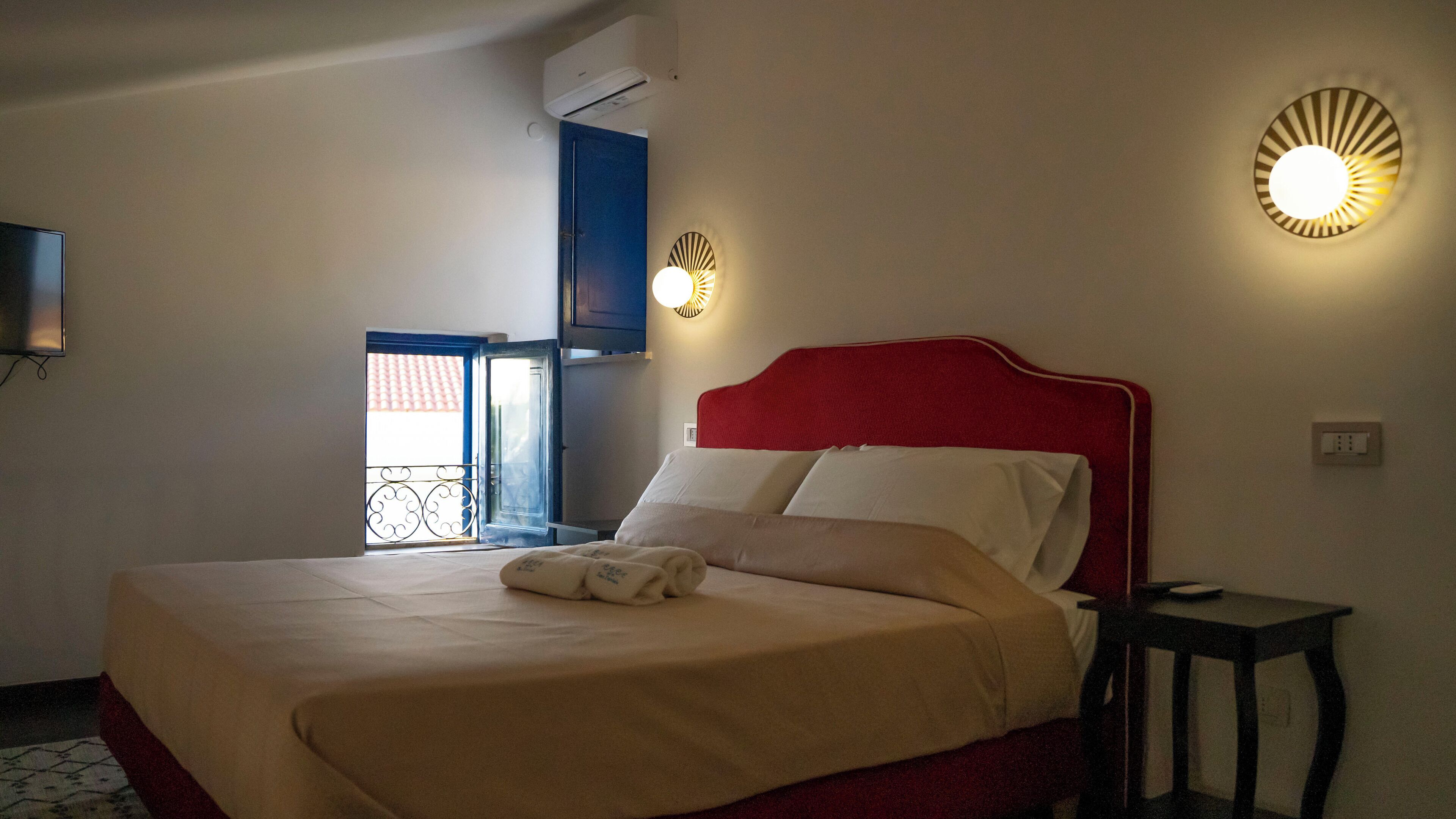 A hotel room with a bed, a bedside table, a wall-mounted light, and a door leading to a balcony.