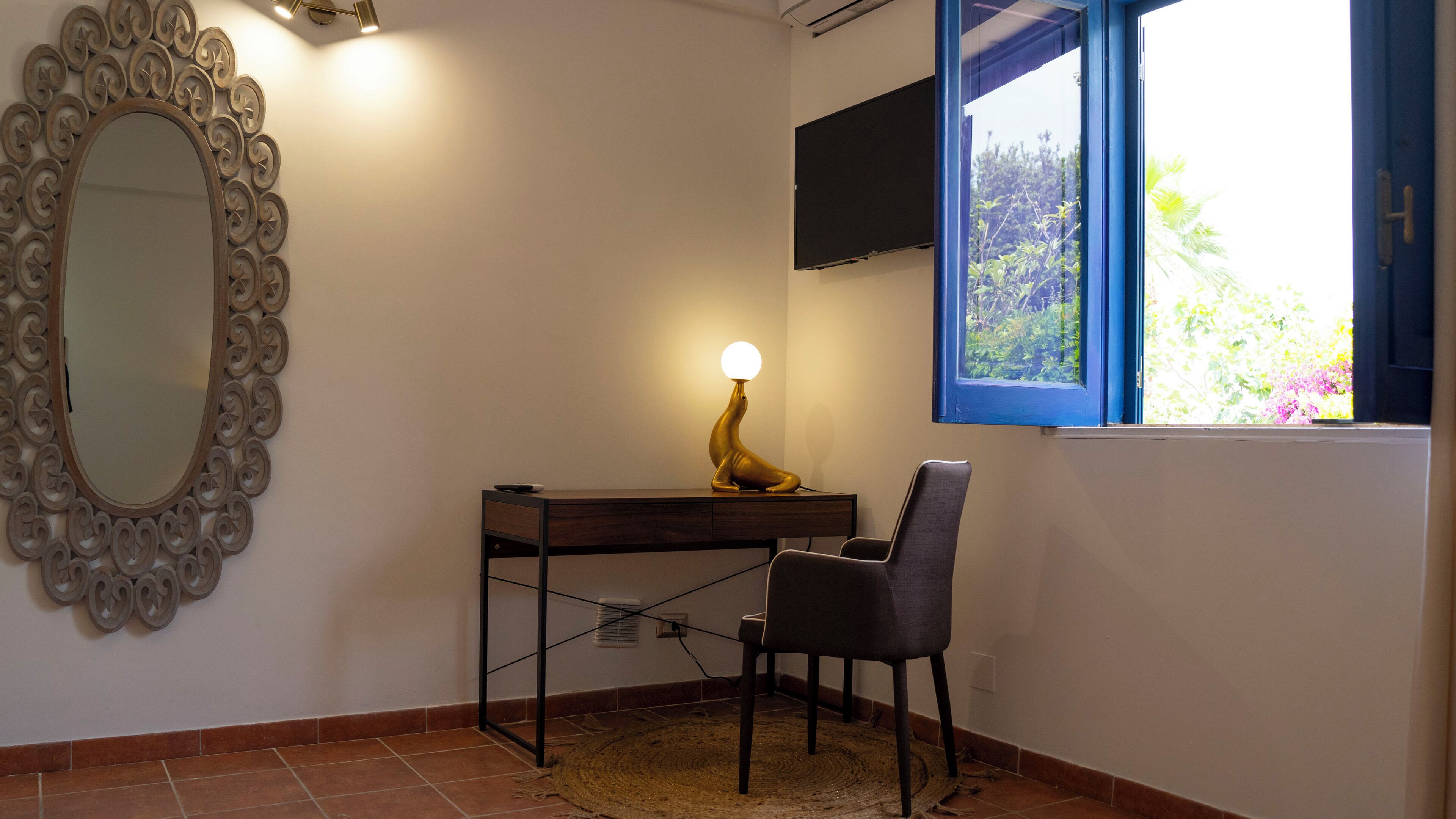 Una habitación con escritorio, silla, espejo decorativo y ventana con vista a follaje.
