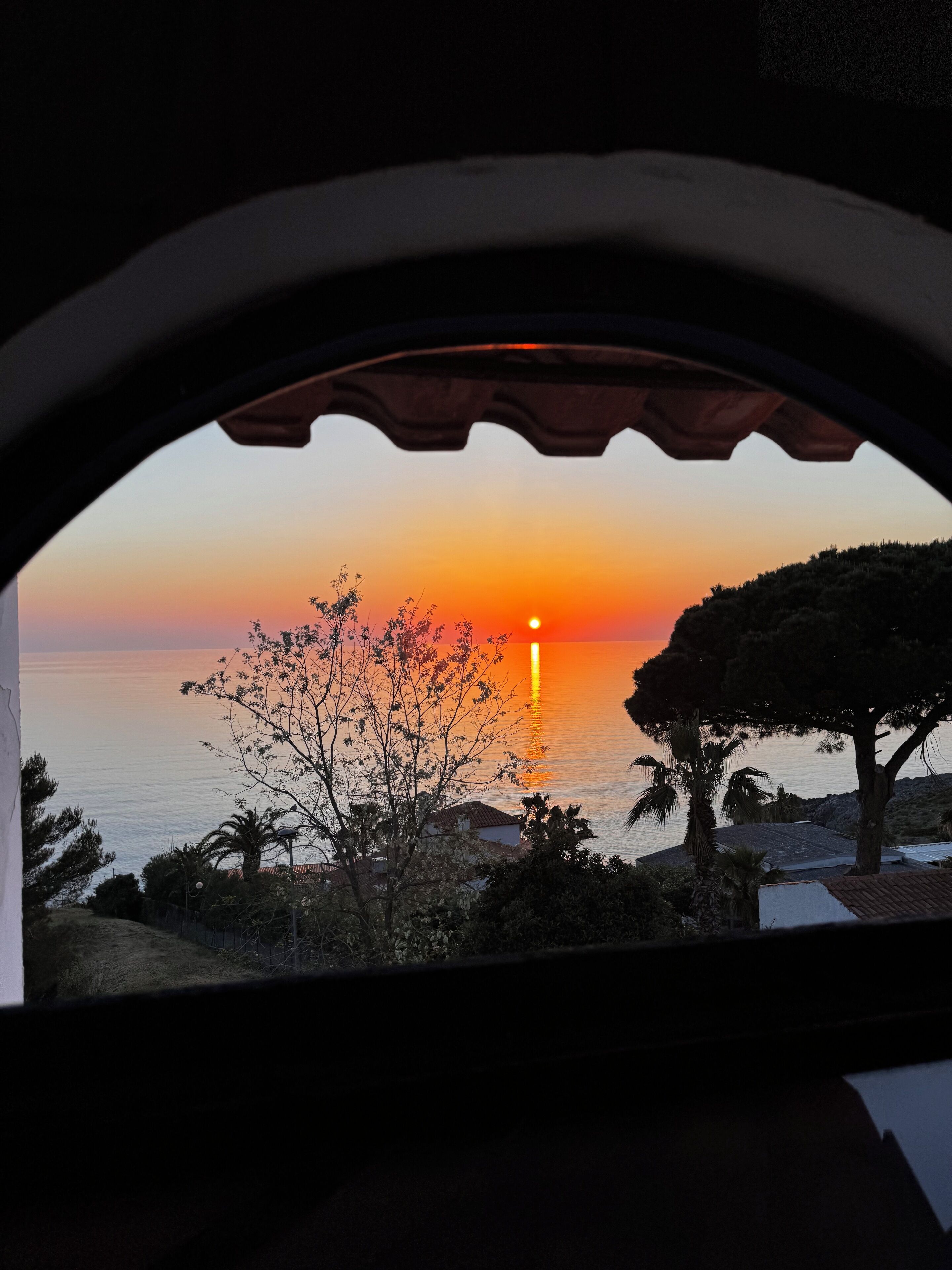 A sunset view through a window with a balcony, overlooking the sea and some trees.