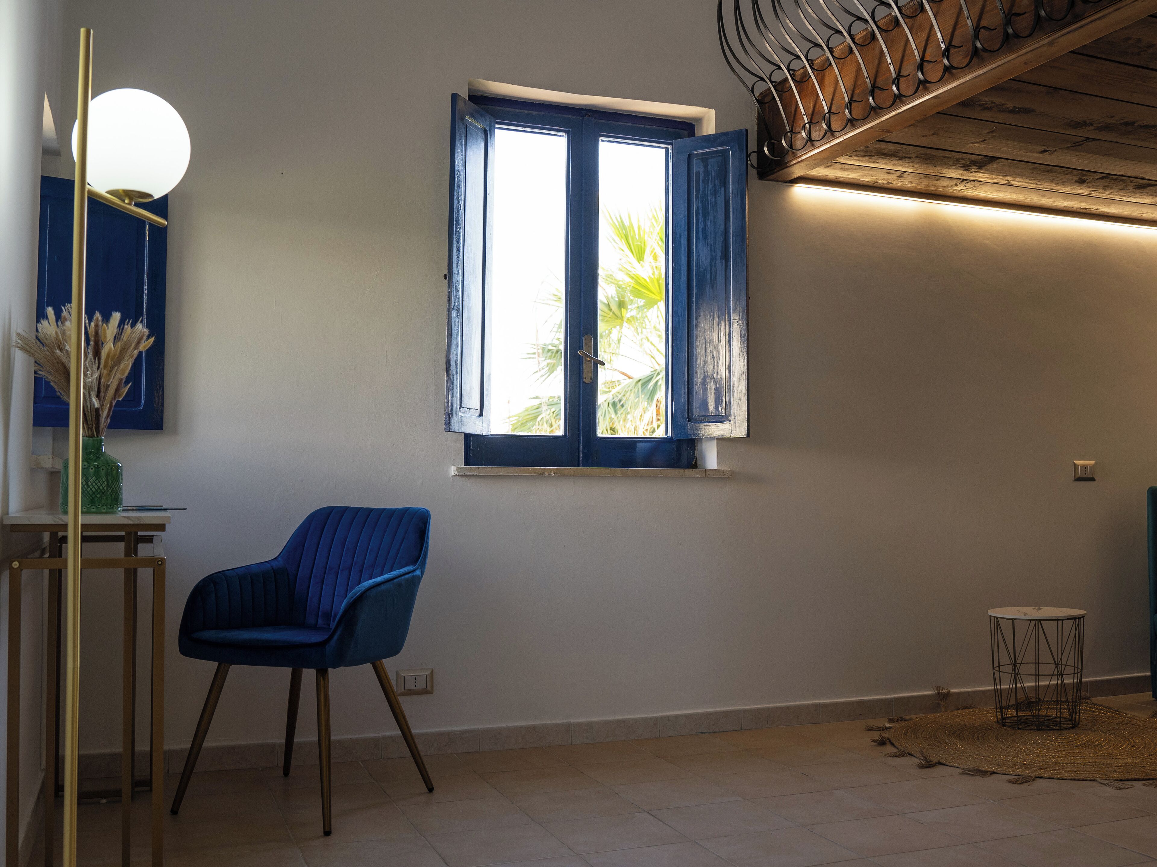 Una habitación con una silla azul, una mesita pequeña, una lámpara de pared y un ventanal con contraventanas azules.