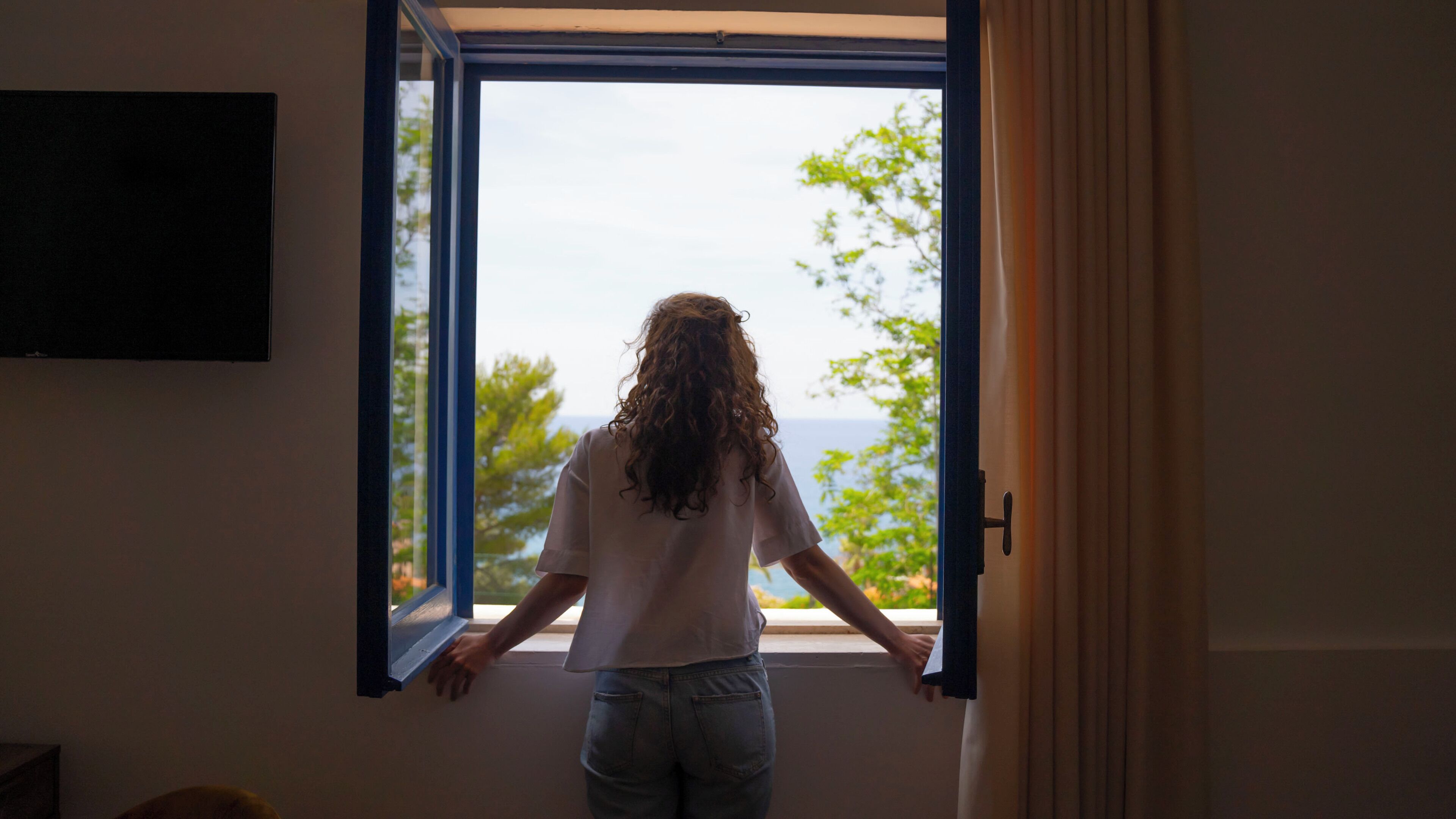 A person standing by an open window with a view of trees and a body of water.
