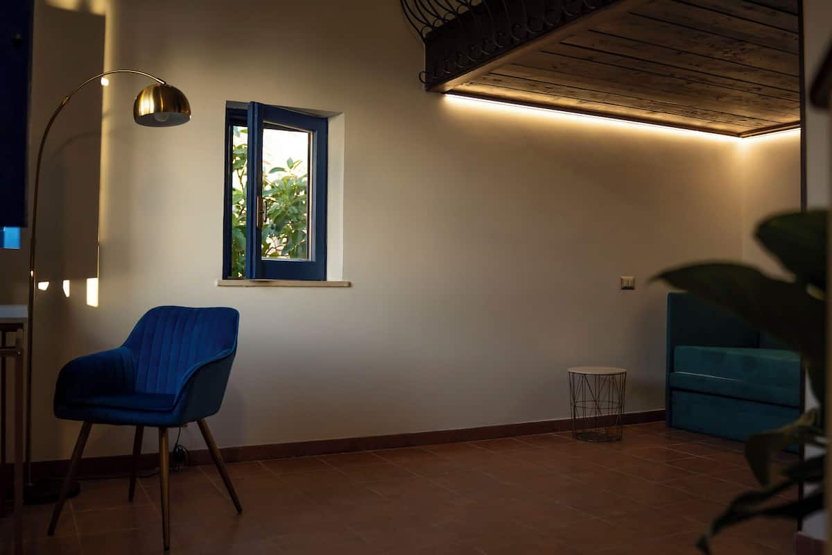 Een kamer met een blauwe stoel, een klein rond tafeltje, een raam met uitzicht op groen en een plafondlamp.