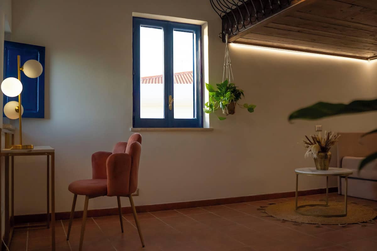 Een kamer met een blauwe deur, een raam met uitzicht, een hangende plant, een kleine ronde tafel en een stoel.