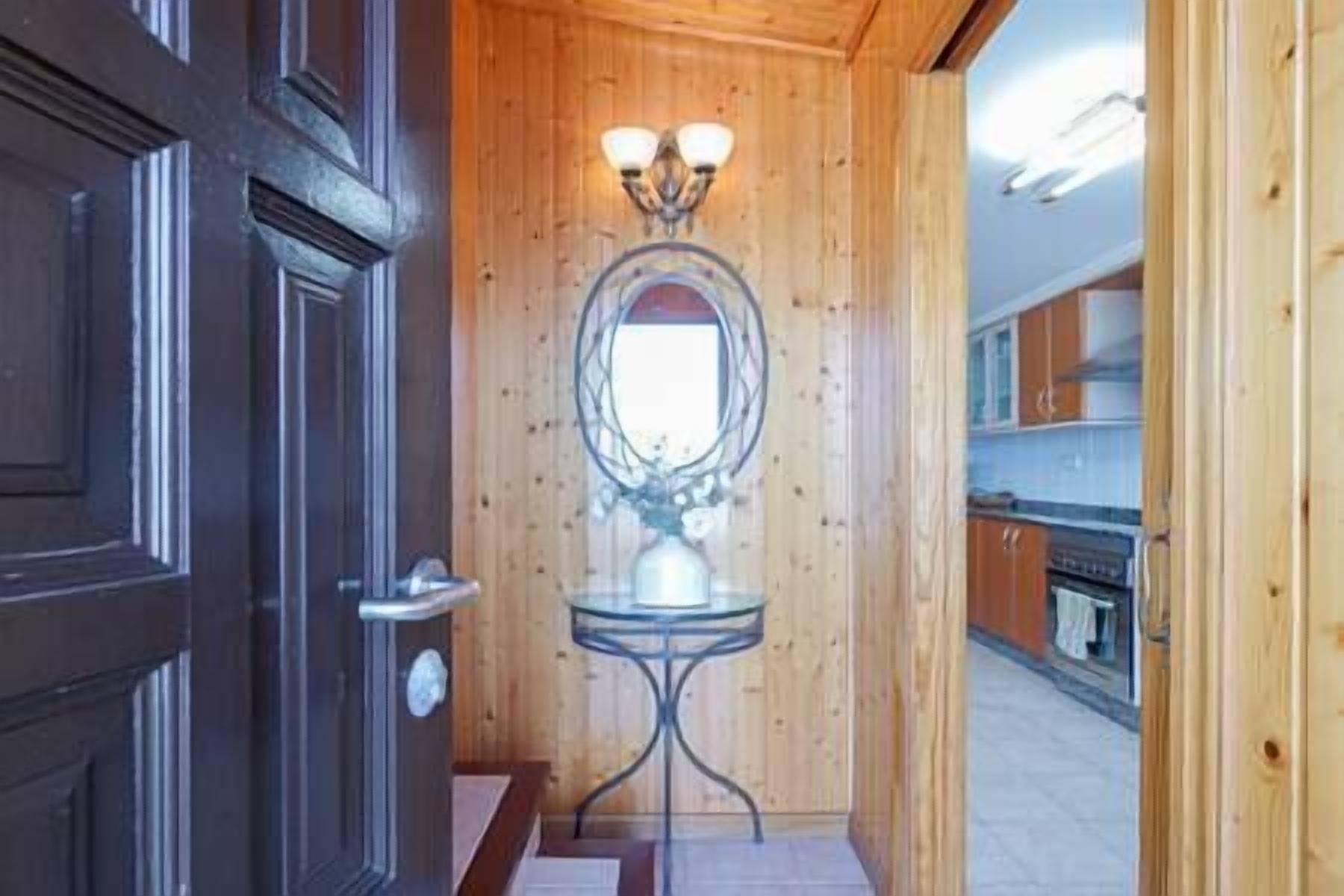 A wooden interior hallway with a door, a mirror with lights, and a glimpse of a kitchen area.