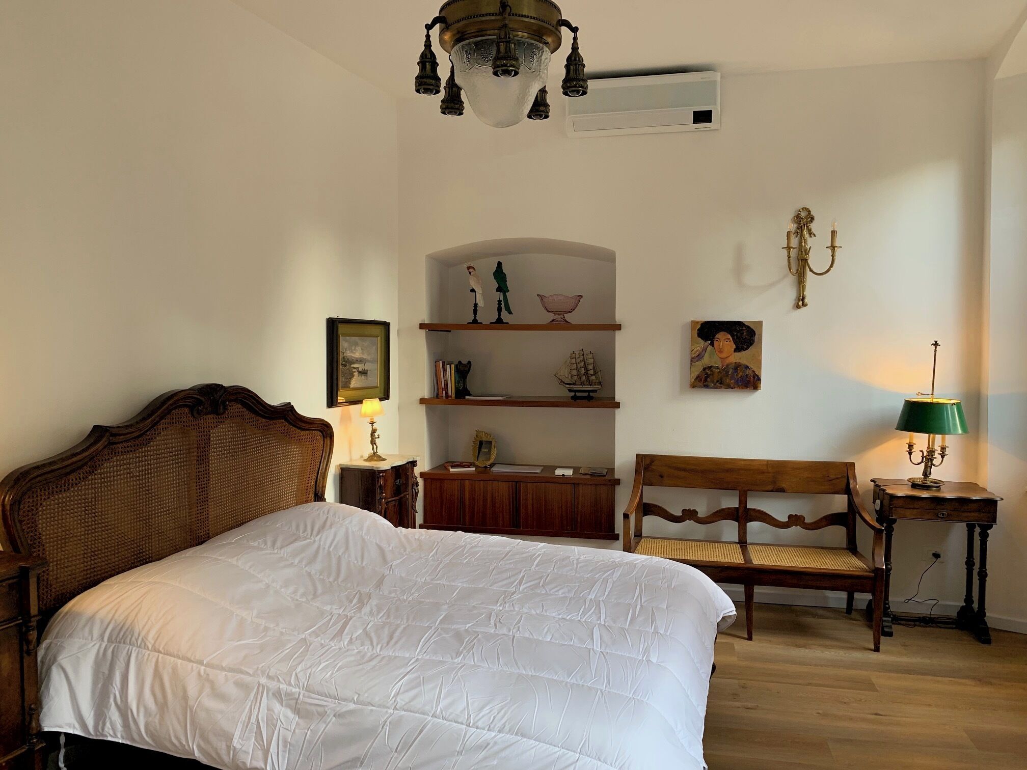 Een slaapkamer met een groot bed, een houten bankje, een kleine tafel met een lamp en wandplanken met decoratieve objecten.