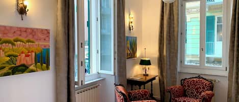 A room with a large window, two armchairs, a table with a lamp, and a painting on the wall.