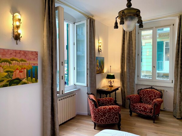 A room with a large window, two armchairs, a table with a lamp, and a painting on the wall.