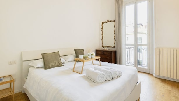 A neatly made bed with rolled towels, a bedside table with a tray, a mirror, and a window with a view of buildings. - Easylife - Modern and central apt in Porta Romana (Milano)