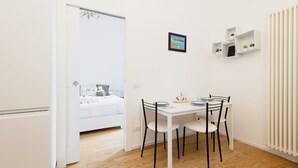 A small, modern room with a white dining table and chairs, a bed with white bedding, and a radiator on the wall. - Easylife - Modern and central apt in Porta Romana (Milano)